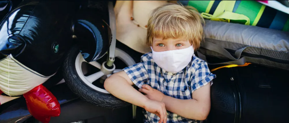 Foto de un niño con mascarilla sentando en el maletero de un coche lleno de cosas