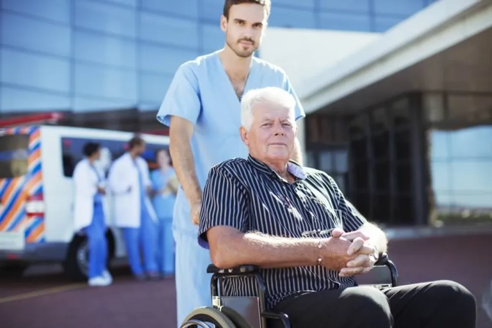 Hombre mayor en silla de ruedas acompañado por un profesional hospitalario en la entrada del Hospital